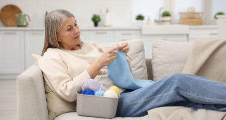 Senior woman knitting with needles on sofa at home