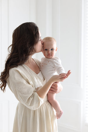 Mother in dress kissing her cute baby near white wall at home