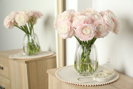 Beautiful ranunculus flowers in glass vase and jewelry on wooden chest of drawers near white wall...