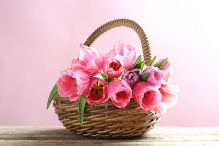 Beautiful flowers in wicker basket on wooden table against pink backgroundの写真素材