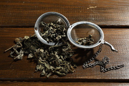 Metal infuser with dry tea leaves on wooden table, closeup