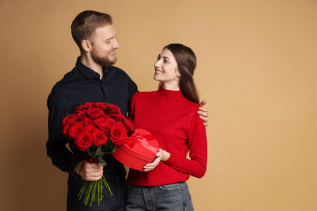 Happy couple with bouquet of red roses and gift box on beige background, space for text