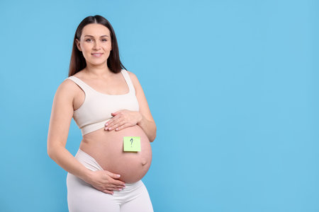 Choosing name for baby. Pregnant woman with note with question mark on her belly against light blue background, space for textの写真素材