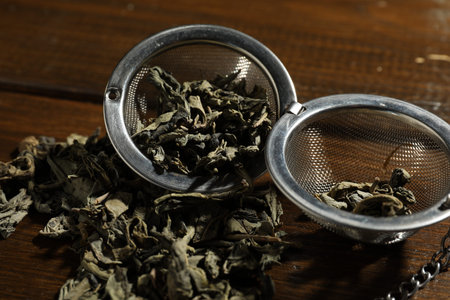 Metal infuser with dry tea leaves on wooden table, closeup