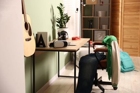 Wooden table with laptop, decor, chair, clothes and guitar in stylish teenager's room