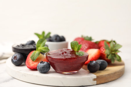Different sweet jams in glass bowls, strawberries, blueberries and mint on white marble table,...