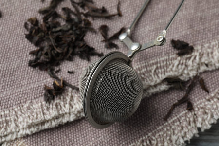 Metal infuser and dry tea leaves on grey cloth, closeup