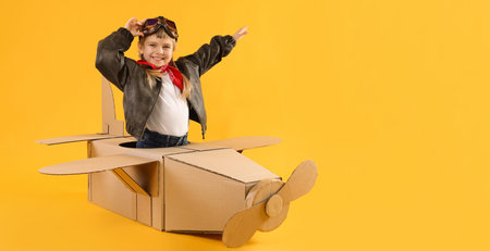 Happy little girl in cardboard airplane, goggles, jacket and handkerchief playing pilot on yellow...