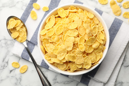Corn flakes in bowl and spoon on white marble table, flat lay