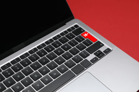 Laptop with 18 plus button on red background, closeup. Mature content warning