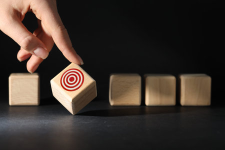 Woman touching wooden cube with target among other ones against black background, closeup