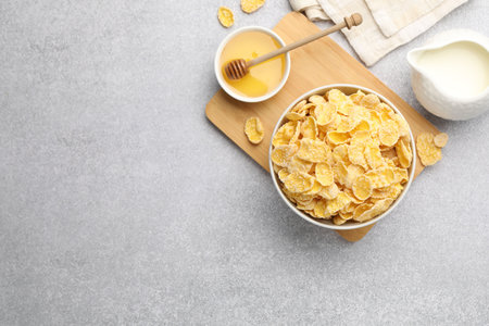 Tasty corn flakes in bowl, milk and honey on light grey background, flat lay. Space for text