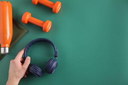 Man with stylish headphones, dumbbells and thermo bottle on green background, top view. Space for...