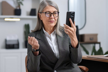 Beautiful senior businesswoman in suit and glasses using smartphone on chair in office