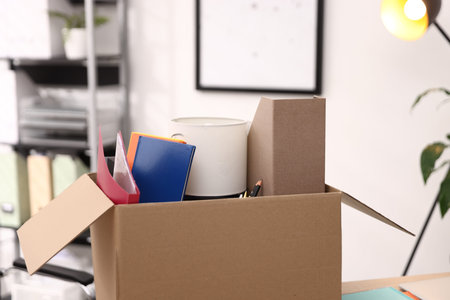 Different personal belongings in cardboard box on table in office, closeup