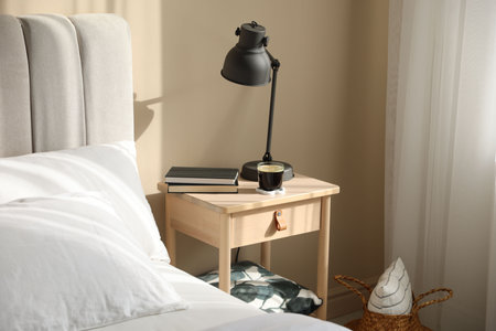 Stylish lamp, books and cup of coffee on wooden bedside table near bed indoors