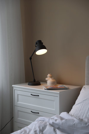 Stylish lamp and magazines on white dresser near bed indoors