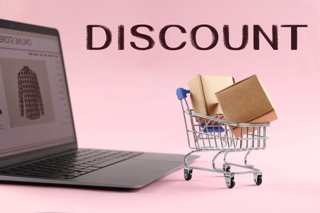 Small shopping cart with cardboard boxes and laptop on pink background