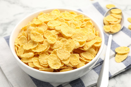 Tasty corn flakes in bowl and spoon on white marble table, closeup
