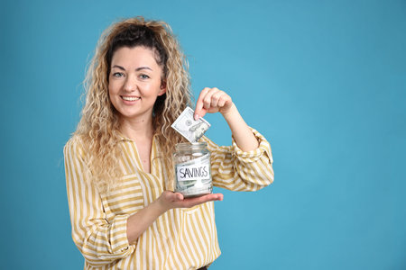 Happy woman with savings jar on light blue background, space for text