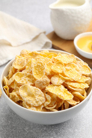 Tasty corn flakes in bowl, milk and honey on light grey background, closeup