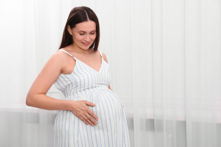 Beautiful pregnant woman touching her belly near curtains at home, space for text