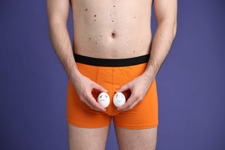 Urology. Man in orange underwear holding eggs with drawn unhappy faces near his genitals on purple...
