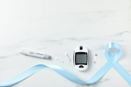 Diabetes. Digital glucometer, pen, test strip, lancets and light blue ribbon on white marble table,...