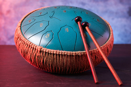 Steel tongue drum and mallets in red light on dark table near textured wall, closeup