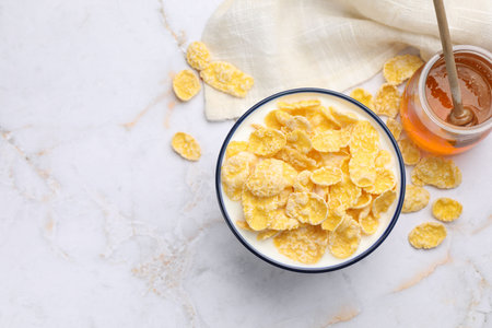 Tasty corn flakes in bowl served with honey on white marble table, flat lay. Space for text