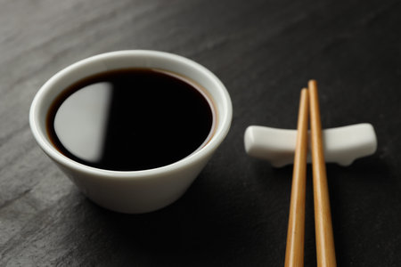 Soy sauce in bowl and chopsticks on black table, closeup