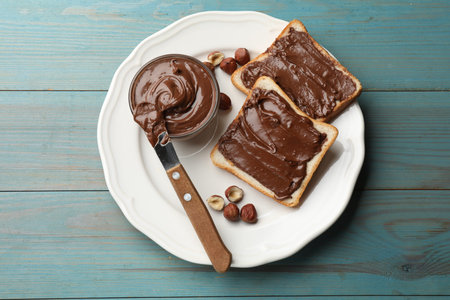 Toasts with chocolate paste, hazelnuts and knife on light blue wooden table, top view