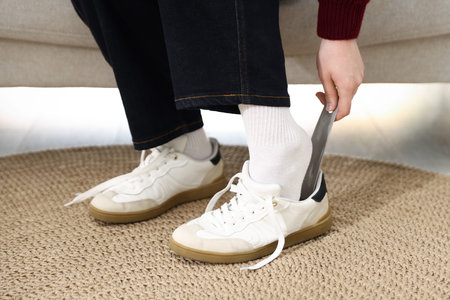 Woman putting on stylish sneakers with shoehorn indoors, closeup