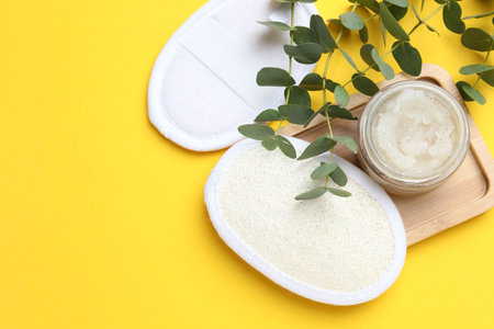 Natural loofah sponges, scrub and eucalyptus branches on yellow background, flat lay. Space for text