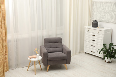 Stylish armchair, side table, chest of drawers, decor and houseplant near window indoors. Interior...