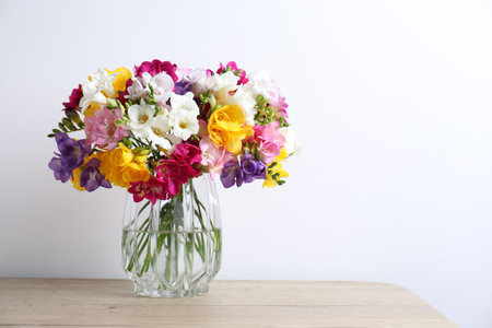 Beautiful freesia flowers in glass vase on wooden table near white wall indoors, space for textの写真素材