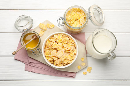 Tasty corn flakes in bowl served with honey and milk on white wooden table, flat lay