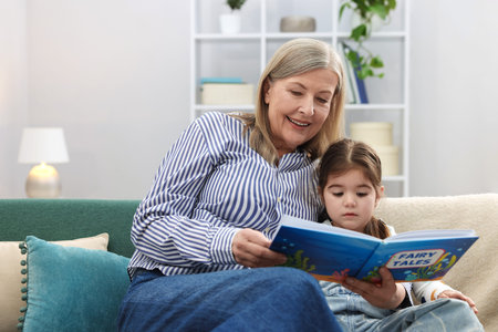 Nanny reading book to cute little girl on sofa indoorsの写真素材
