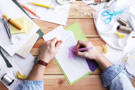 Fashion designer drawing sketch of outfit at wooden desk, top view