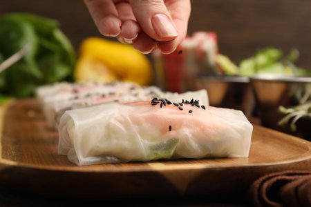 Woman sprinkling fresh spring rolls wrapped in rice paper with black sesame seeds against blurred...