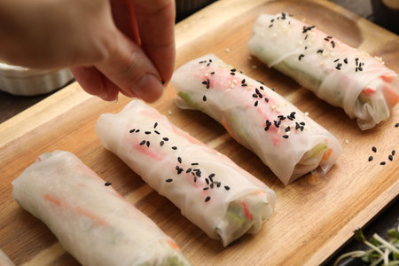 Woman sprinkling fresh spring rolls wrapped in rice paper with sesame seeds at table, closeup