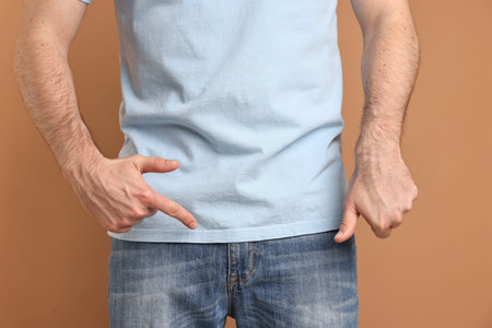 Urology. Man pointing at his groin and showing thumbs down on beige background, closeup