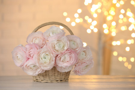 Many beautiful ranunculus flowers in wicker basket on light wooden table against blurred lights,...