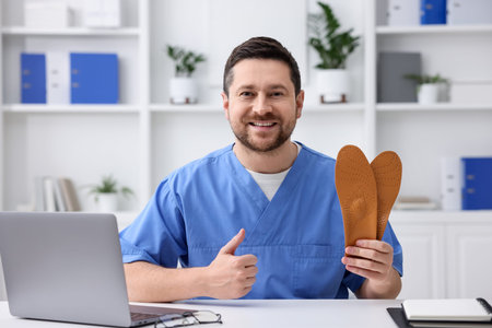 Smiling orthopedist in scrubs with foot insoles showing thumbs up at white desk in clinicの写真素材