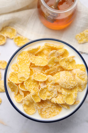 Tasty corn flakes in bowl served with honey on white marble table, flat lay