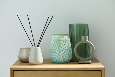 Beautiful vases, reed air freshener and candle holder on wooden bedside table near white wall...