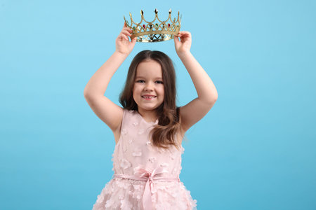 Beautiful little girl with crown dreaming about becoming queen on light blue backgroundの写真素材