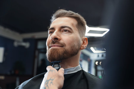 Stylist trimming man's beard with hair clipper in barbershop, closeup