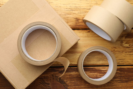 Eco-friendly packaging. Rolls of masking tape and cardboard box on wooden table, flat lay