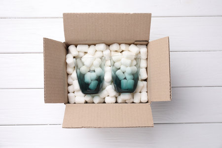 Cardboard gift box with styrofoam pieces and glasses on white wooden table, top view